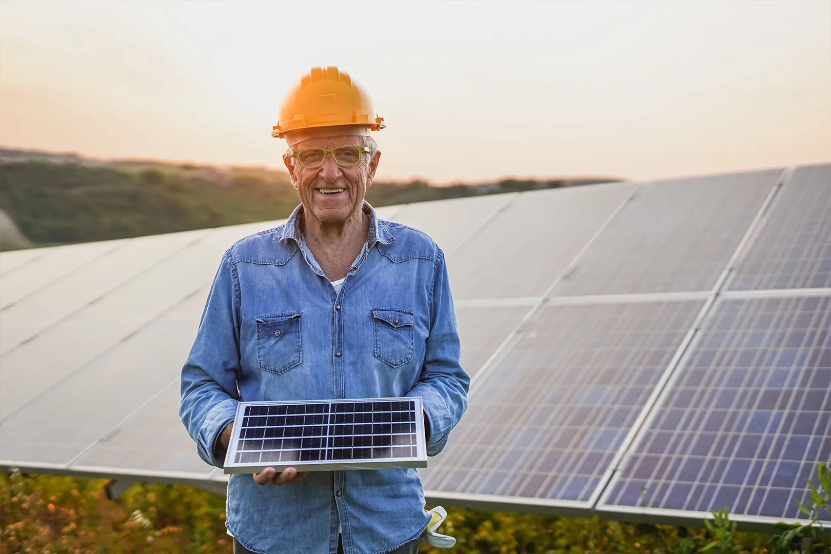 Älterer Mann mit Bauhelm hält ein Solarmodul vor einem Solarpark – Experte im Ruhestand leitet ein Bauvorhaben mit Sentaris